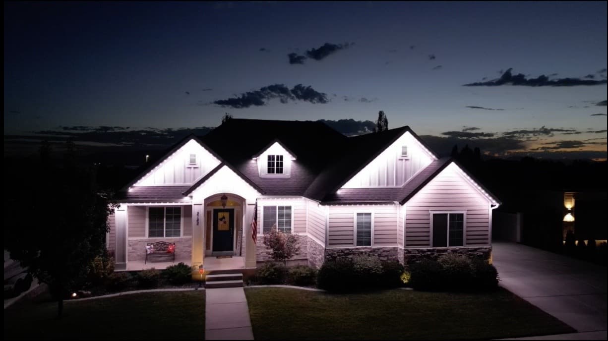 Light-colored single-story home with stone accents and bright white permanent lighting on several front-facing gables.