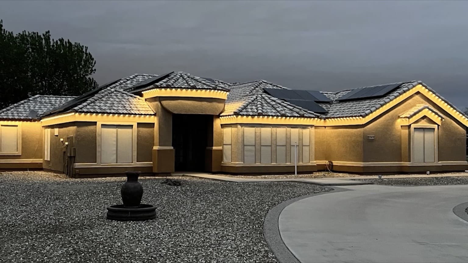 Single-story stucco home with tile roof and warm-white permanent lighting following long horizontal eaves and entry rooflines.