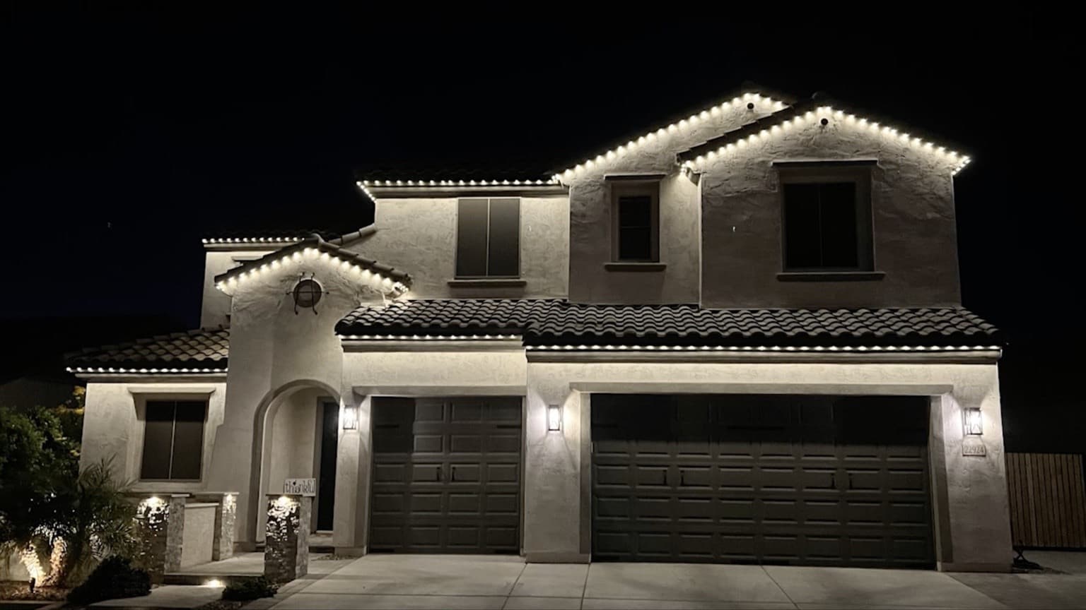 Two-story stucco home with tile roof and warm-white permanent lighting outlining layered gables and the long garage eave.